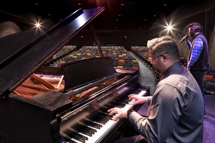 A man plays a grand piano on stage while another person sings into a microphone. They are performing in an empty auditorium with rows of colorful seats and bright stage lights shining above.
